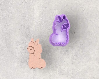 May include: Two llama-shaped cookie cutters. One is a light peach color, and the other is purple. The llama shapes have a fluffy body and a smiling face. The purple cutter has a scalloped edge.