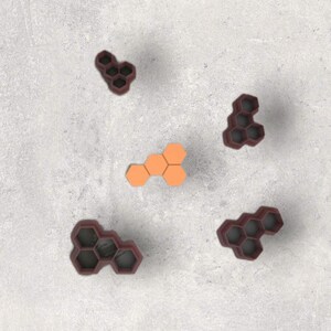 May include: An overhead shot shows several hexagon-shaped cookie cutters in shades of orange and maroon. The cutters are scattered on a light gray surface. The cutters are arranged in clusters of three and five, with one cluster of orange hexagons.