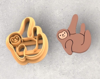 May include: A light orange plastic sloth-shaped cookie cutter next to a brown sloth-shaped decoration. The cookie cutter has a detailed outline of a sloth hanging from a branch. The brown decoration has a smiling face.