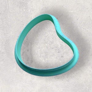 Puede incluir: Un cortador de galletas abstracto de color turquesa con forma de óvalo curvo e irregular. El cortador tiene un borde liso y redondeado y un ancho constante. El objeto está sobre un fondo gris claro texturizado.