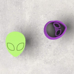 Puede incluir: Dos cortadores de galletas con temática alienígena. Uno es una cabeza de alienígena verde lima con ojos ovalados. El otro es un cortador de galletas con forma de cabeza de alienígena púrpura con la misma forma de ojos ovalados.
