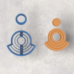 May include: Two geometric cookie cutters. One is blue and features a circular design with concentric rings and a separate small circle. The other is orange and has a similar design, with a small circle above a larger, layered shape.