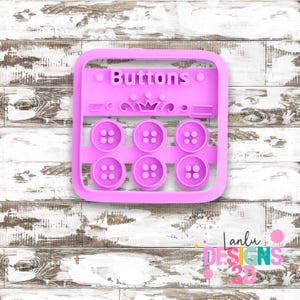 May include: A square, light purple plastic mold featuring the word Buttons in raised letters at the top, a decorative crown icon in the center, and six circular button shapes arranged in two rows of three, each with four holes in the middle.