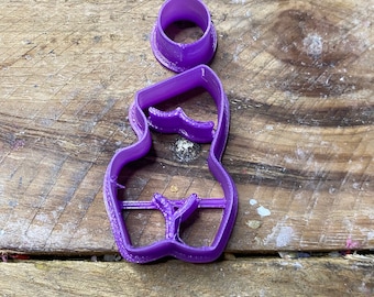 Può includere: Un tagliabiscotti viola a forma di corpo femminile, con un pezzo separato a forma di anello. Il tagliabiscotti è realizzato in plastica e si trova su una superficie di legno. Il design include dettagli del corpo.