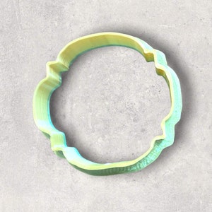 May include: A 3D-printed cookie cutter in a circular shape with a scalloped edge. The cutter is a gradient of yellow and light blue, with a textured appearance. The design is suitable for baking and crafting.