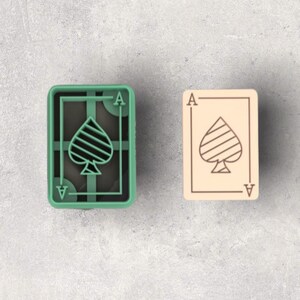 Puede incluir: Un cortador de galletas verde con forma de As de Picas junto a una galleta beige con forma de As de Picas. El cortador de galletas tiene un diseño de pica con la letra "A" en cada esquina. La galleta tiene el mismo diseño de pica y la letra "A" en cada esquina.