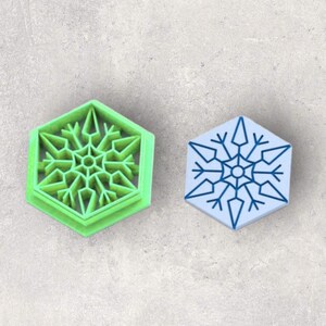 May include: A bright green hexagonal cookie cutter featuring a detailed snowflake design with a central point and radiating arms, positioned next to a light blue hexagonal stamp showing the same snowflake pattern in a darker blue ink.