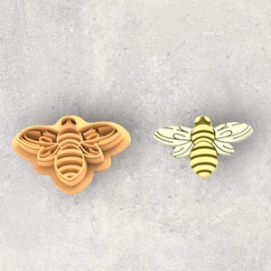 May include: Two bee-shaped cookie cutters, one is a light brown color and the other is a light yellow color. Both cutters have a detailed design of a bee with wings, a body, and legs.