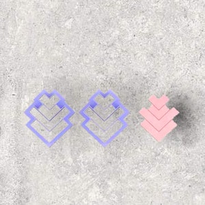 May include: Three geometric shaped cookie cutters. Two are light purple, and the third is pink. Each cutter has a layered, chevron-like design. The cutters are arranged on a gray, textured surface.