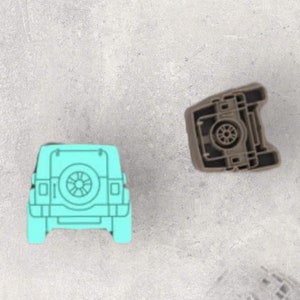 May include: A light turquoise plastic cookie cutter shaped like the rear view of an off-road vehicle with a spare tire mounted on the back, positioned next to a brown plastic stamp piece that features a detailed internal mechanical structure.