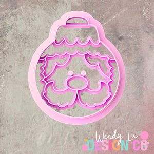May include: A pink Christmas ornament-shaped cookie cutter featuring a Santa Claus face design. The cutter has a round shape with a handle at the top and a detailed face with a mustache, beard, and nose. The text "Wendy Lu Design Co" is visible.