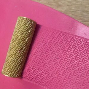 May include: A gold-coloured rolling pin with a geometric pattern sits on a pink, textured surface. The pattern is also imprinted on the pink surface, creating a matching design. The items appear to be for crafting or baking.