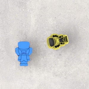 May include: Two cookie cutters: one blue, shaped like a toy soldier with a moustache, and one yellow, shaped like a bell. The cutters are on a grey surface. The toy soldier is standing upright.