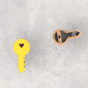 May include: Two key-shaped objects with heart cutouts. One is yellow with a dark purple heart, the other is a light brown color. The keys are on a light gray surface. The keys are likely decorative or for crafts.