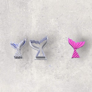 May include: Three mermaid tail cookie cutters are shown. One is light blue and grey, another is a light blue outline, and the third is bright pink. The cookie cutters are on a grey surface.