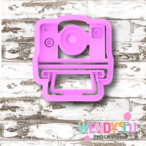 May include: A pink plastic cookie cutter shaped like a vintage instant camera. The cutter features a rectangular body, a circular lens, and a slot for the photo. The design includes details such as the flash and viewfinder.