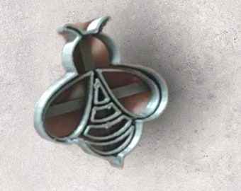 Può includere: Un tagliabiscotti a forma di ape color argento. Il tagliabiscotti ha un design dettagliato con un corpo arrotondato, ali e antenne. Il corpo dell'ape presenta diverse linee curve e il tagliabiscotti è tenuto su uno sfondo neutro.