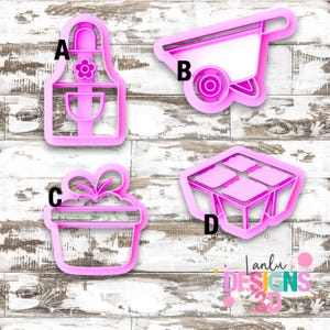 Peut inclure: Quatre emporte-pièces en plastique rose étiquetés A, B, C et D. A est un tablier, B une brouette, C un coffret cadeau avec un nœud, et D une jardinière à quatre sections. Les objets sont disposés sur une surface en bois blanc avec un logo.