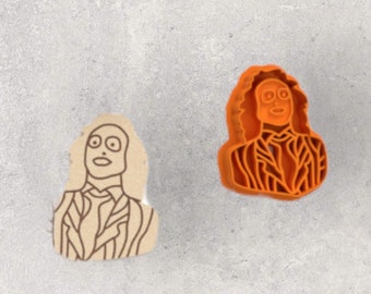 May include: Two cookie cutters in the shape of a character with wild hair and a striped suit. One is a tan cookie with a black outline, and the other is an orange plastic cutter.