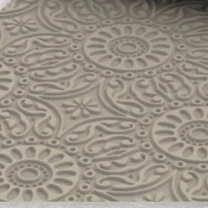 Op de afbeelding: Close-up van een beige, getextureerd oppervlak met een herhalend patroon van gestileerde bloemen en cirkelvormige ontwerpen. Het patroon is in reliëf, waardoor een verhoogd reliëfeffect ontstaat. Het ontwerp omvat bloemen, cirkels en wervelende lijnen.