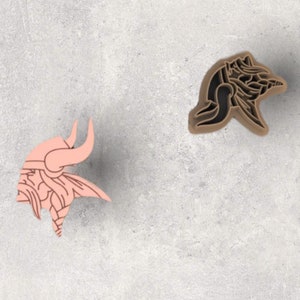 May include: Two Viking-themed cookie cutters. One is a light pink Viking head with a horned helmet. The other is a brown bear-shaped cutter. Both cutters are on a gray surface.