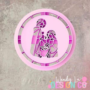 May include: A pink, circular design featuring a religious scene with figures, likely a nativity or holy family depiction. The design is layered, with a detailed cutout effect. The outer ring has a segmented pattern. The image includes the text "Wendy Lu Design Co."