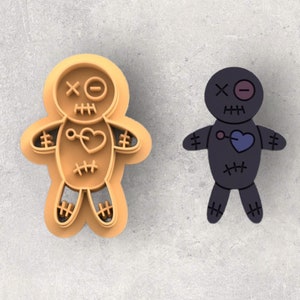 May include: A voodoo doll cookie cutter and a voodoo doll illustration. The cookie cutter is tan and shaped like a doll with stitched details, a heart, and a cross for an eye. The illustration is a dark purple doll with a blue heart.
