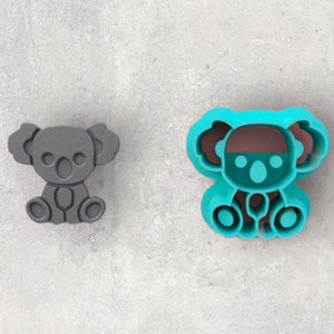 May include: Two koala-shaped cookie cutters. One is solid gray, the other is teal with brown and teal accents. The koalas are sitting with their legs together and arms at their sides. The cutters are made of plastic and are on a gray surface.