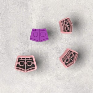 May include: Four cookie cutters in the shape of denim shorts. One purple cutter is solid, while the other three are pink with black interior detailing. The cutters are arranged on a gray surface.