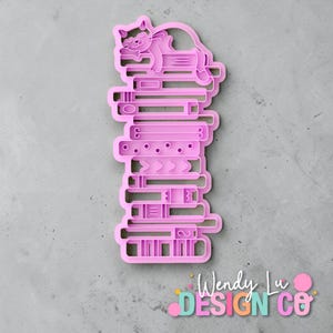 May include: A pink cookie cutter in the shape of a stack of books with a cat sleeping on top. The cookie cutter has detailed designs on the books, including lines, circles, and arrows. The text "Wendy Lu Design Co" is at the bottom.