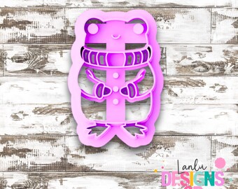May include: A light pink plastic cookie cutter shaped like a hamster wearing a scarf. The design features a rounded body, small ears, a striped scarf around the neck, and tiny paws held near the center. The cutter has a thick, raised edge.
