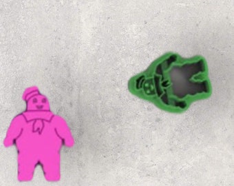 May include: A pink, cartoon-like figure of a marshmallow man with a hat and scarf, and a green cookie cutter in the shape of a ghost. The objects are on a gray surface.