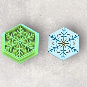 May include: A lime green hexagonal cookie cutter featuring a raised snowflake design and a light blue hexagonal stamp with a dark blue and brown snowflake pattern printed on the surface, both displaying intricate geometric snowflake details.