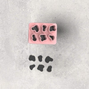 May include: A pink silicone mould with six cow spot shapes and six detached black cow spot shapes. The mould is rectangular with rounded corners. The cow spot shapes are irregular and organic.