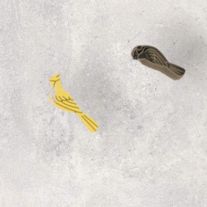 May include: Two wooden bird figurines. One is yellow with light yellow stripes, and the other is brown with darker brown stripes. The birds are positioned on a light grey background.