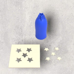 May include: A blue bottle stopper with a star-shaped top, a cream-colored square stencil with star cutouts, and several small star-shaped decorations. The items are arranged on a gray surface.