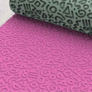 Könnte beinhalten: Eine rosa Teigrolle mit einem sich wiederholenden Muster aus geometrischen Formen, Zahlen und Symbolen. Ein Teil einer grauen Teigrolle mit ähnlichem Design ist im Hintergrund zu sehen. Das Design ist geprägt.