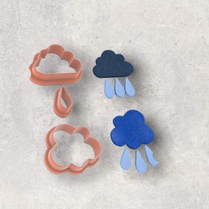 May include: A collection of cloud and raindrop-shaped cookie cutters in a light orange color, alongside two blue cloud-shaped leather embellishments with light blue raindrop cutouts. The items are arranged on a gray surface.
