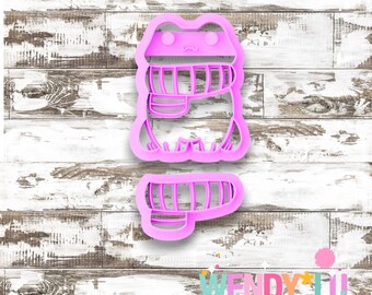 May include: A pink frog-shaped cookie cutter with a detailed design, including a mouth and eyes. The cutter is made of plastic and is designed for cutting out cookies or other baked goods. The design includes the text "WENDY LU DESIGN STUDIO".