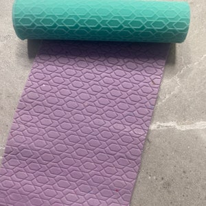 May include: A teal rolling pin with a honeycomb pattern rests above a lavender sheet with the same pattern. The rolling pin is cylindrical, and the sheet is rectangular. The objects are likely for baking or crafting.