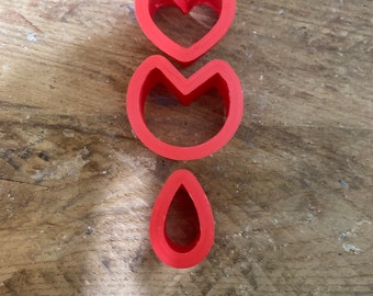 Può includere: Tre formine rosse per biscotti a forma di cuore, cuore stilizzato e goccia. Le formine sono realizzate in materiale flessibile e sono disposte verticalmente su una superficie di legno.