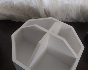 Puede incluir: Un organizador de madera blanca con forma octogonal y cuatro compartimentos internos divididos por paredes que se cruzan. La estructura presenta líneas limpias y un acabado mate suave, proporcionando un espacio segmentado para el almacenamiento.
