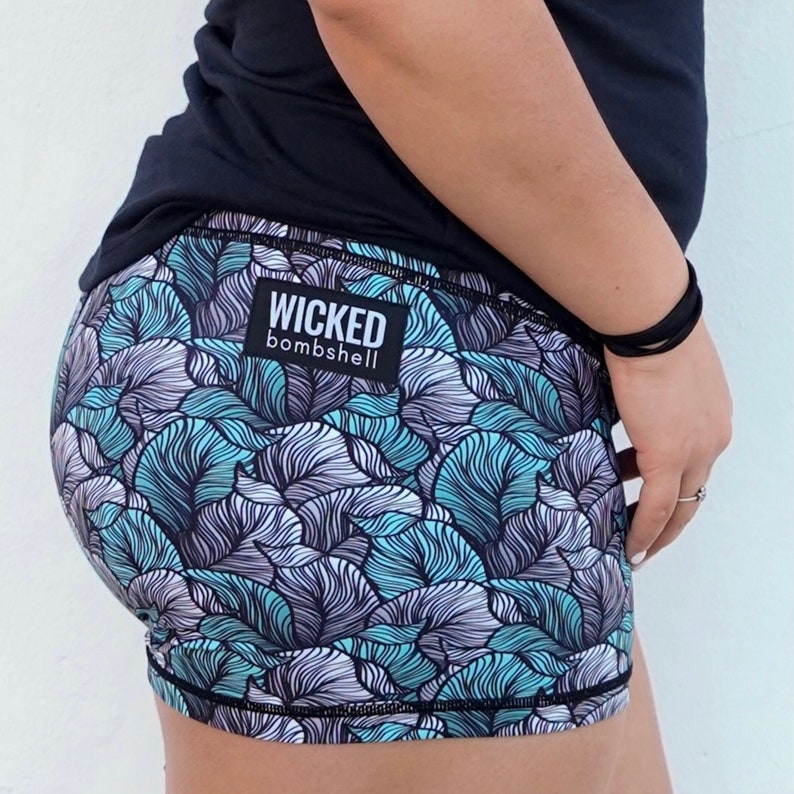 May include: Black athletic shorts with a teal and white floral pattern. The shorts have a black waistband with the text "WICKED bombshell" in white.