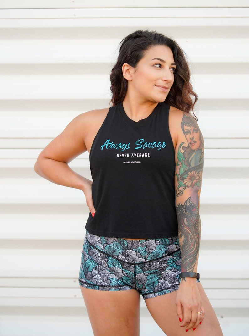 May include: A woman wearing a black tank top with the text "Always Savage Never Average" and floral print athletic shorts. She has a colorful tattoo on her arm.