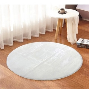 May include: A round, white, fluffy rug sits on a hardwood floor. The rug is the focal point, with a soft, textured appearance. A small white table with a coffee cup and books are in the background, near a sheer white curtain.