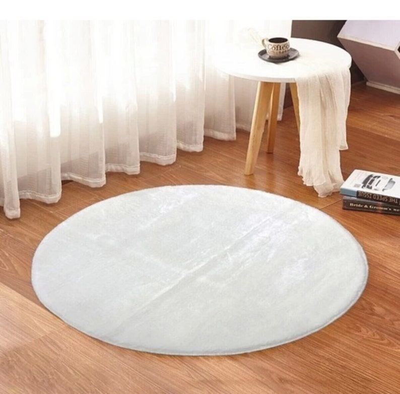 May include: A round, white, fluffy rug sits on a hardwood floor. The rug is the focal point, with a soft, textured appearance. A small white table with a coffee cup and books are in the background, near a sheer white curtain.