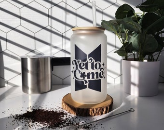 May include: Frosted glass tumbler with a bamboo lid and straw. The tumbler features the text "Yet to Come" in a black graphic design. It sits on a wooden coaster, with a cleaning brush and coffee grounds nearby. A plant and a stainless steel container are in the background.