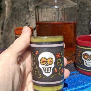 May include: A set of four colorful shot glasses with a black skull and floral design. The glasses are made of ceramic and have a glossy finish.