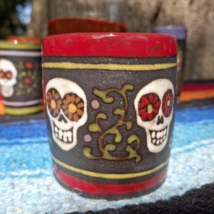 May include: A red ceramic mug with a black background and two white sugar skulls with colorful flowers. The skulls have black eyes and a black outline. The mug has a gold trim around the top and bottom.