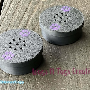 May include: Two gray, round containers with a textured surface and a series of small holes in the center. Each container is decorated with purple paw prints. The containers are sitting on a wooden surface.